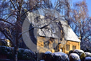 An old English house is all in the snow, next to trees and bushes.