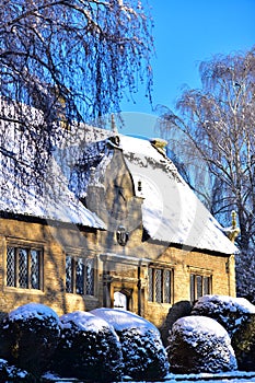 An old English house is all in the snow, next to trees and bushes.