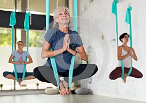 Old European female balancing in Lotus Pose Padmasana in blue hammock during fly yoga in fitness club