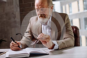 Old exhausted man is putting his glasses on the desk while making notes