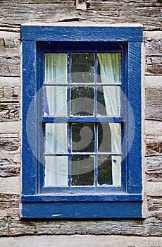 Old faded light wood building with a dark blue wooden window frame and white curtains pulled back