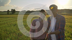 Old farmer discussing new project with his successor and holding tablet, standing on wheat field during beautiful sunset