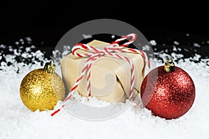 Old fashioned gift wrapped in plain paper, tied with red and white twine sitting in snow beside holiday ornaments