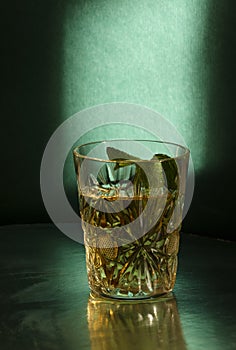 Old-fashioned glass with alcohol drink and piece of mint against green wall. Low and muffled light