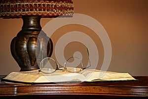 Old fashioned lamp on desk beside open book with glasses laying on it- Room for copy