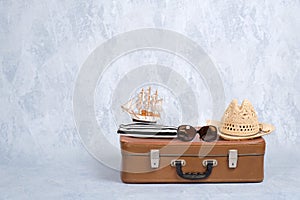 Old fashioned leather travel bag with summer marine accessories: glasses, straw beach hat, toy sailboat on grey background. Banner