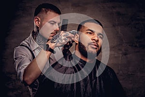 Old-fashioned professional tattooed hairdresser does a haircut to an African American client. on dark textured