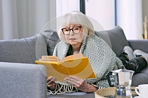 Old fashioned senior woman laying on the couch reading a book.