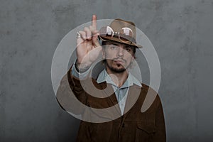 Old-fashioned strong young man in a vintage hat in a round glasses in a stylish brown jacket stands near a gray wall in the studio