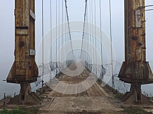Old foggy suspension bridge entrance. Altai village bridge. Early misty morning