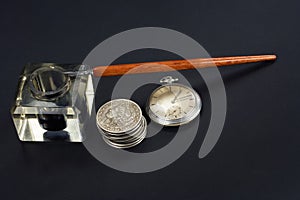 Old fountain pen and inkwell with silver coins and pocket watch on a black background