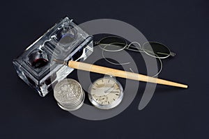 Old fountain pen and inkwell with silver coins and pocket watch on a black background