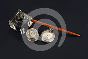 Old fountain pen and inkwell with silver coins and pocket watch on a black background