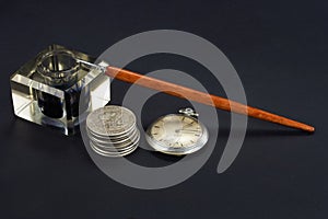 Old fountain pen and inkwell with silver coins and pocket watch on a black background