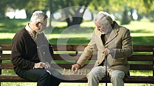 Old friends sitting on bench in park and playing chess, happy leisure time