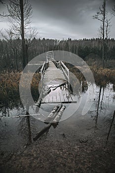 Old and gloomy wooden bridge over the river. Detachment in the water. Gloomy and mysterious forest. Cloudy sky.