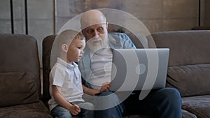 Old grandfather communicates with young grandson, sitting at laptop, watching cartoons, communicating via video chat