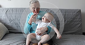 An old grandmother with deep walruses sits on the couch and holds a small grandson in her arms.
