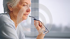 Old grandmother holds glasses in wrinkly hand with serious face sitting against blurred window under sunlight at home