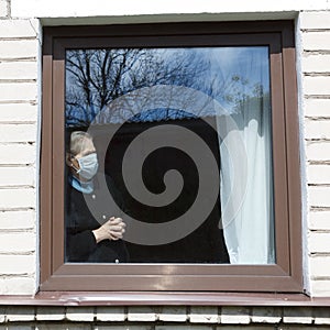 An old grandmother in a protective mask looks out the window at self-isolation.