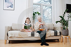 Old granny sitting with her granddaughter on the couch, reading and knitting