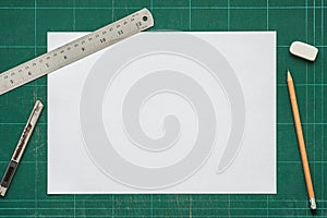 Old green dirty cutting mat, with empty white paper A4 size, stainless steel ruler, cutter knife, pencil and eraser, top view of