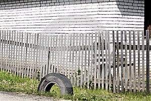 An old grey boardwalk wooden fence with parallel planks and a used tire on the green grass in front of a white brick wall on a