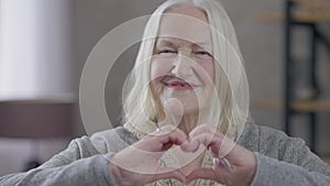 Old grey-haired Caucasian woman showing heart shape with hands looking at camera smiling. Portrait of happy positive