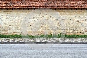 Old grunge yellow brick wall with terracotta tiled roof. A stripe of grass, concrete tiled sidewalk and asphalt road in front. photo