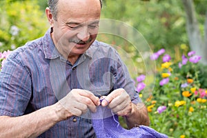 Old handsome man sitting at his summer garden with the knitting needles.