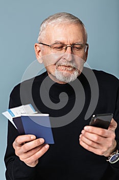 Old handsome smiling man with gray hair and beard in eyeglasses and sweater holding tickets and passports in hand while