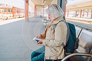 Old happy woman with backpack waiting train on station platform with big bag, passport and ticket on urban background