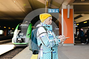 Old happy woman with backpack in yellow hat waiting train on station platform and using smart phone on urban background