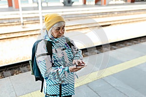 Old happy woman with backpack in yellow hat waiting train on station platform and using smart phone on urban background