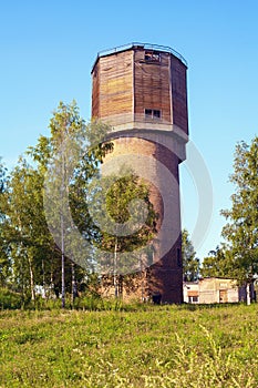 An old high brick water tower with a wooden plank annex on top in the rays of the rising sun among the grass and trees