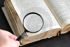 Old historic paper text book opened and magnifying glass in hand. Bible, dictionary, antique document research with loupe.