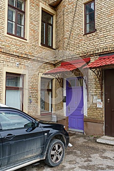 An old historical building made of red brick, a blue wooden door under a canopy and modern plastic double-glazed windows. A parked