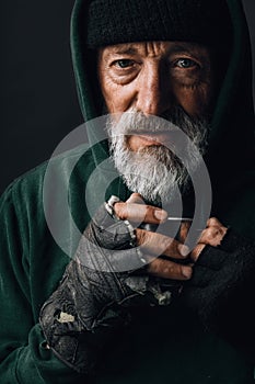 Old homeless man with grey beard holding a mug of hot tea to warm himself.