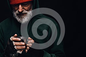 Old homeless man with grey beard holding a mug of hot tea to warm himself.