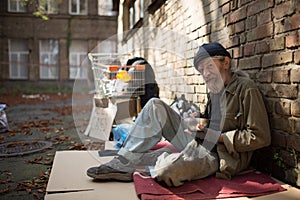 Old homeless man sitting on cardboard holding bowl with food in hand.