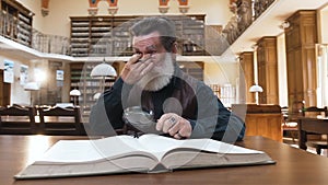 Old intelligent bearded man taking off his glasses and rubbing eyes during his work with book in the library