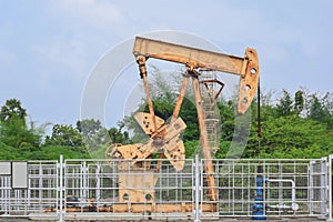 Old isolated rusty oil pump jack extracting crude oil and natural gas form well in green and cloudy oil fields of India, Asia