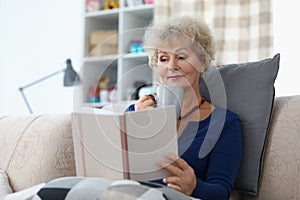 Old lady reading book and drinking cup of coffee being comfy on sofa at home