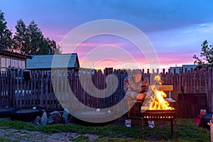 An old lonely man sits sadly on a bench at a village house behind a fence looking into the fire of a barbecue fire