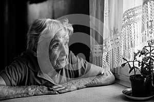 Old lonely woman sitting near the window in his house.