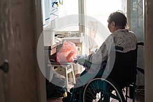 Old lonely woman sitting in a wheelchair near the window in his house