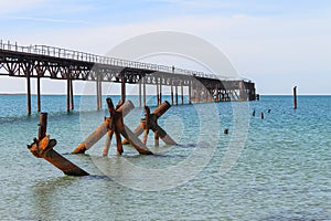 The old long rusty pier going into the sea. There is a man standing on the pier. In the foreground of the water sticking