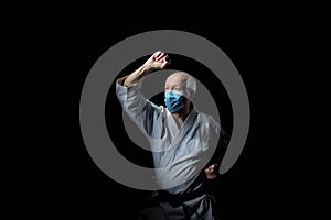 An old male athlete in a medical mask trains a block with his hand on a black background