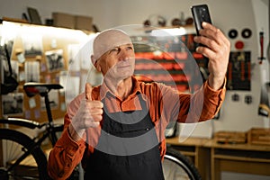 Old male mechanic pausing to take selfie on his mobile phone, repairing or servicing bike in a workshop