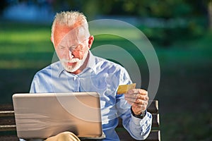 Old male sitting on bench and surfing net on laptop in park, shopping online concept